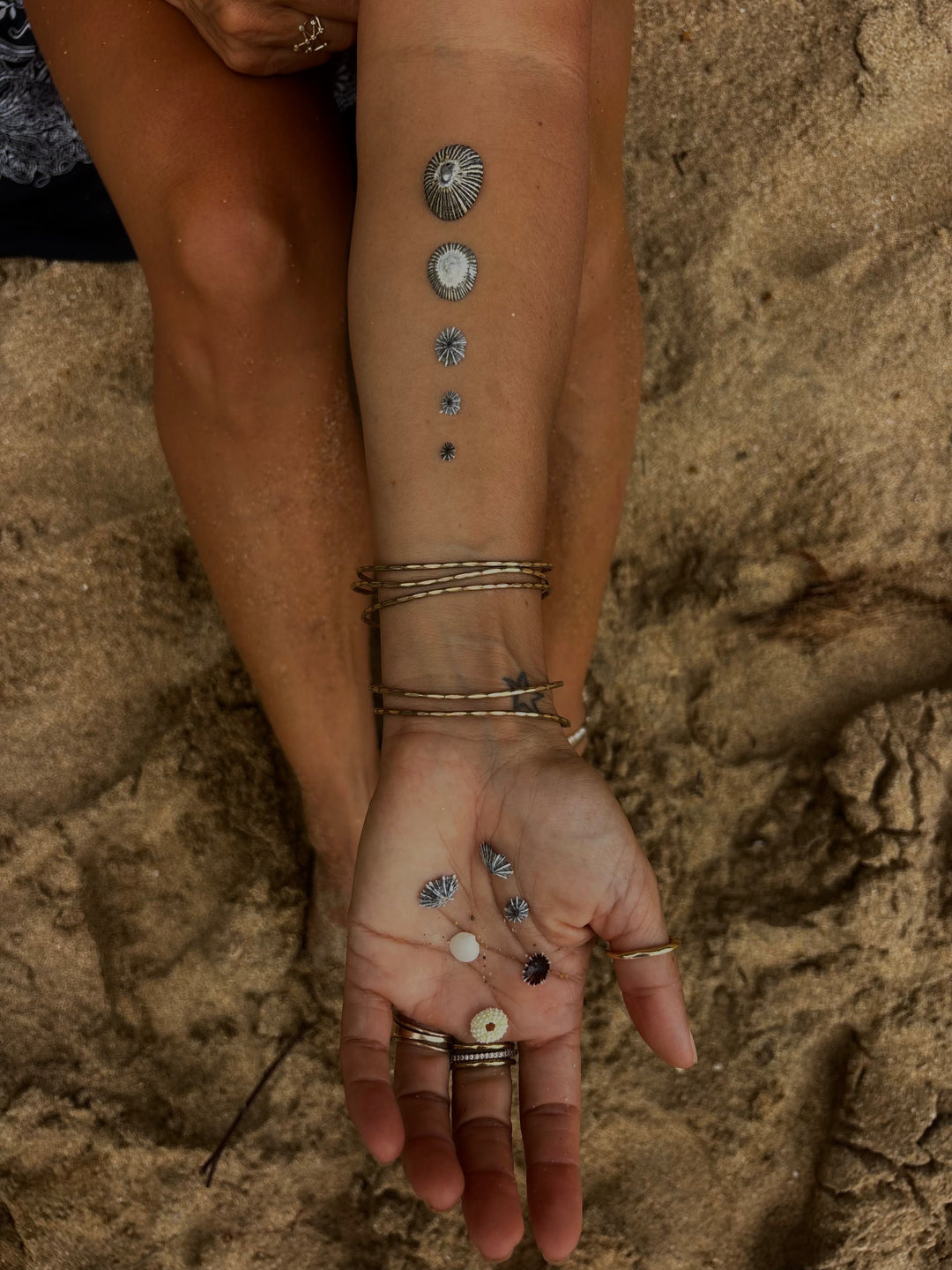 Two bare feet in shallow water on wet sand, stacked silver anklets around one ankle.