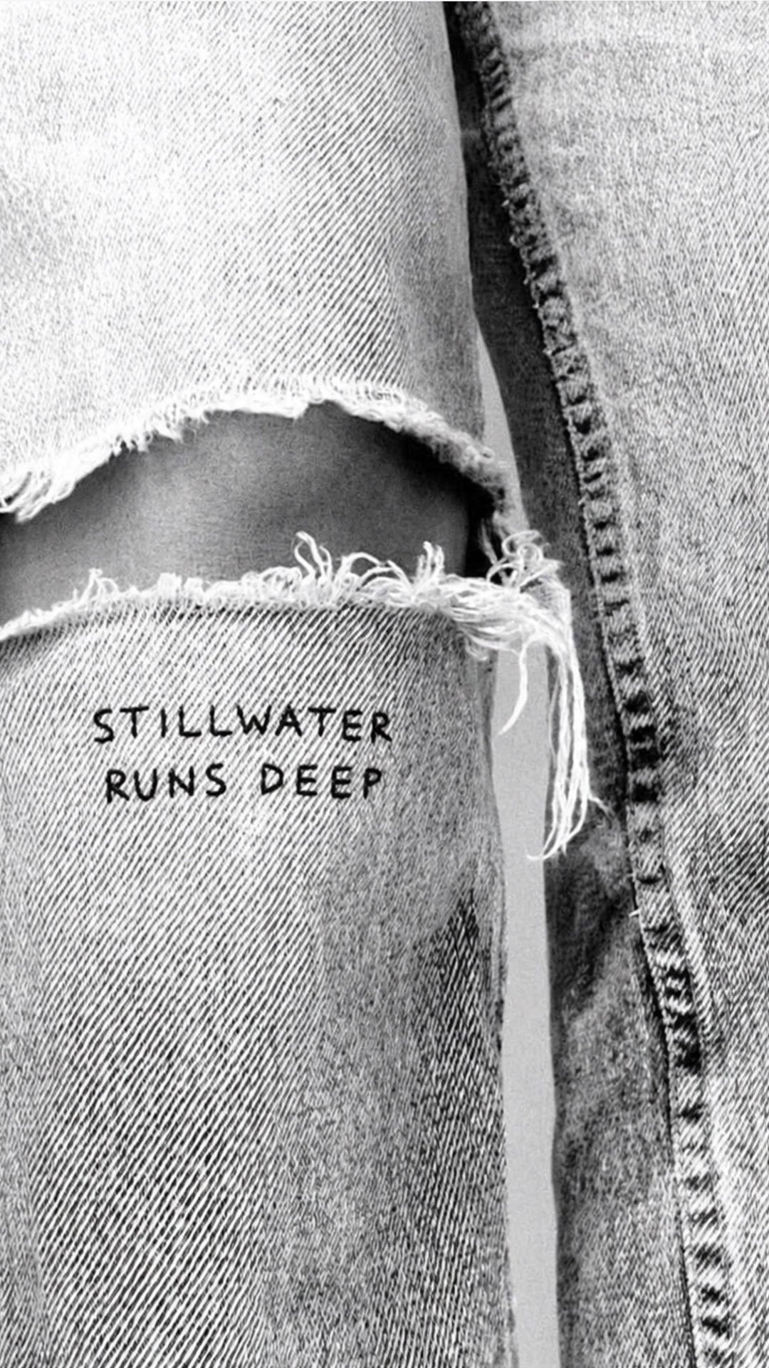 Black and white close-up of denim with a frayed white patch hand-lettered "Stillwater Runs Deep".