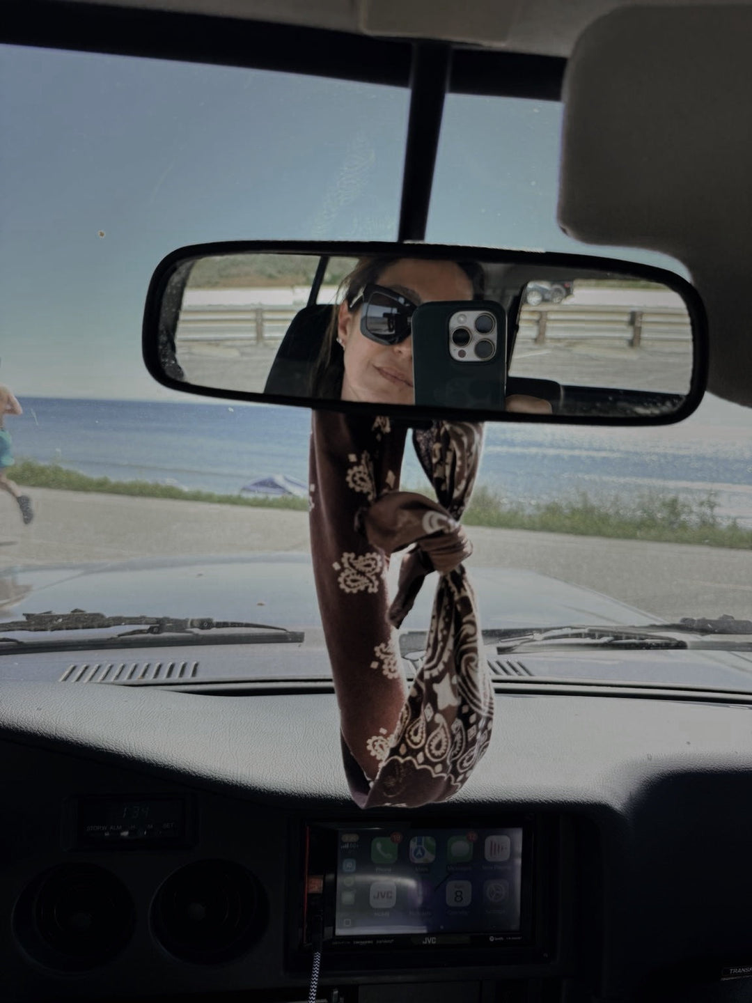 View through a car's bandana clad rearview mirror of a woman photographing her reflection, with the ocean and a parking lot beyond the windshield.