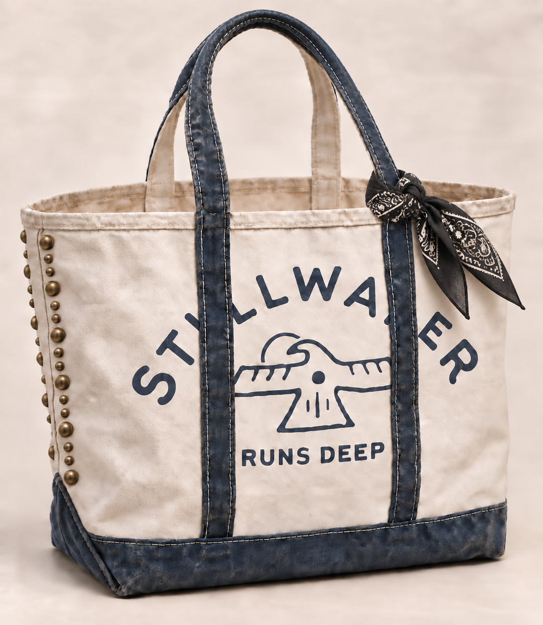 Natural canvas Stillwater tote with brass studs, navy denim straps and a black bandana knotted to one handle, printed "Stillwater Runs Deep" beneath a thunderbird graphic.